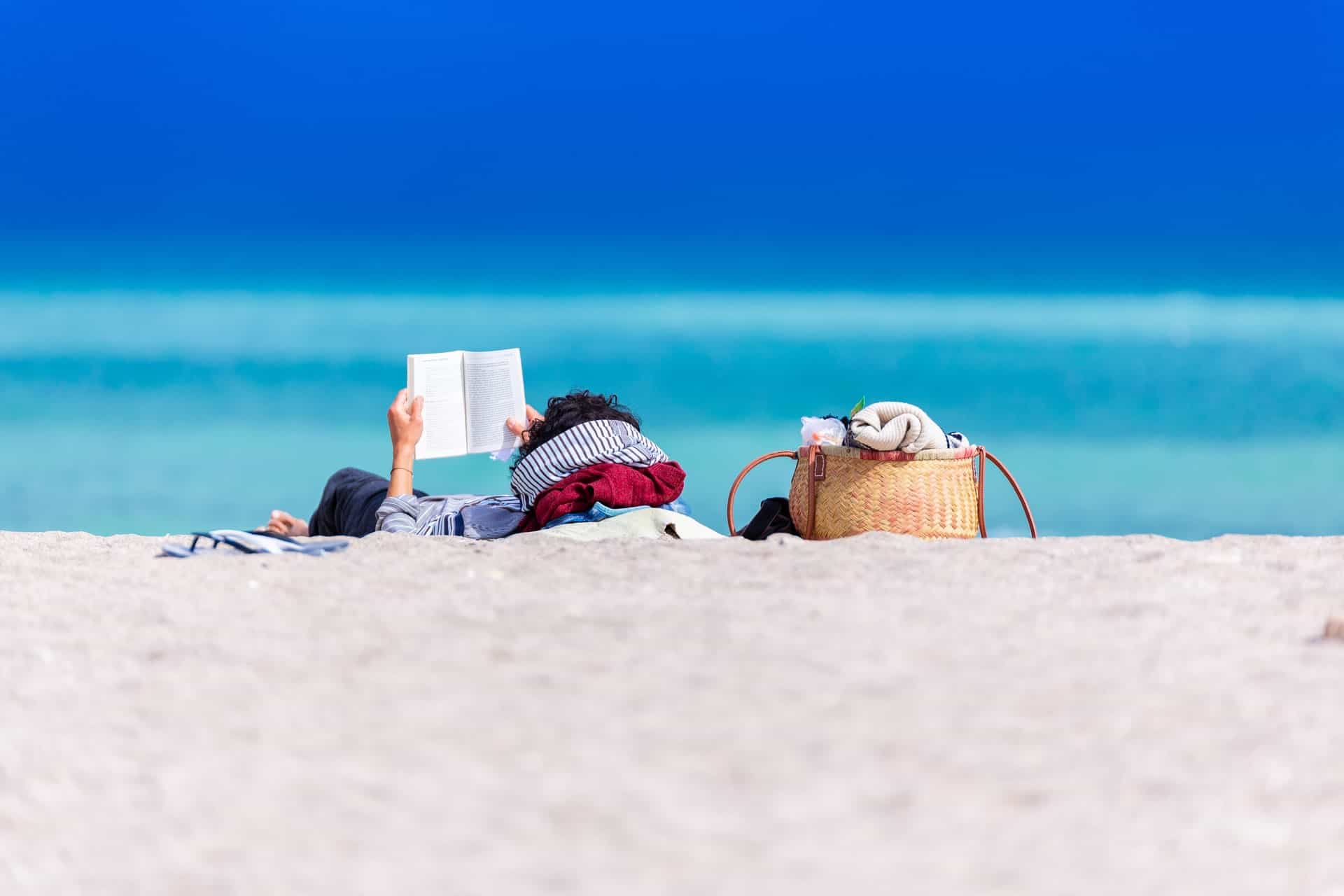 person lying on the shore reading a book next to bags and towels