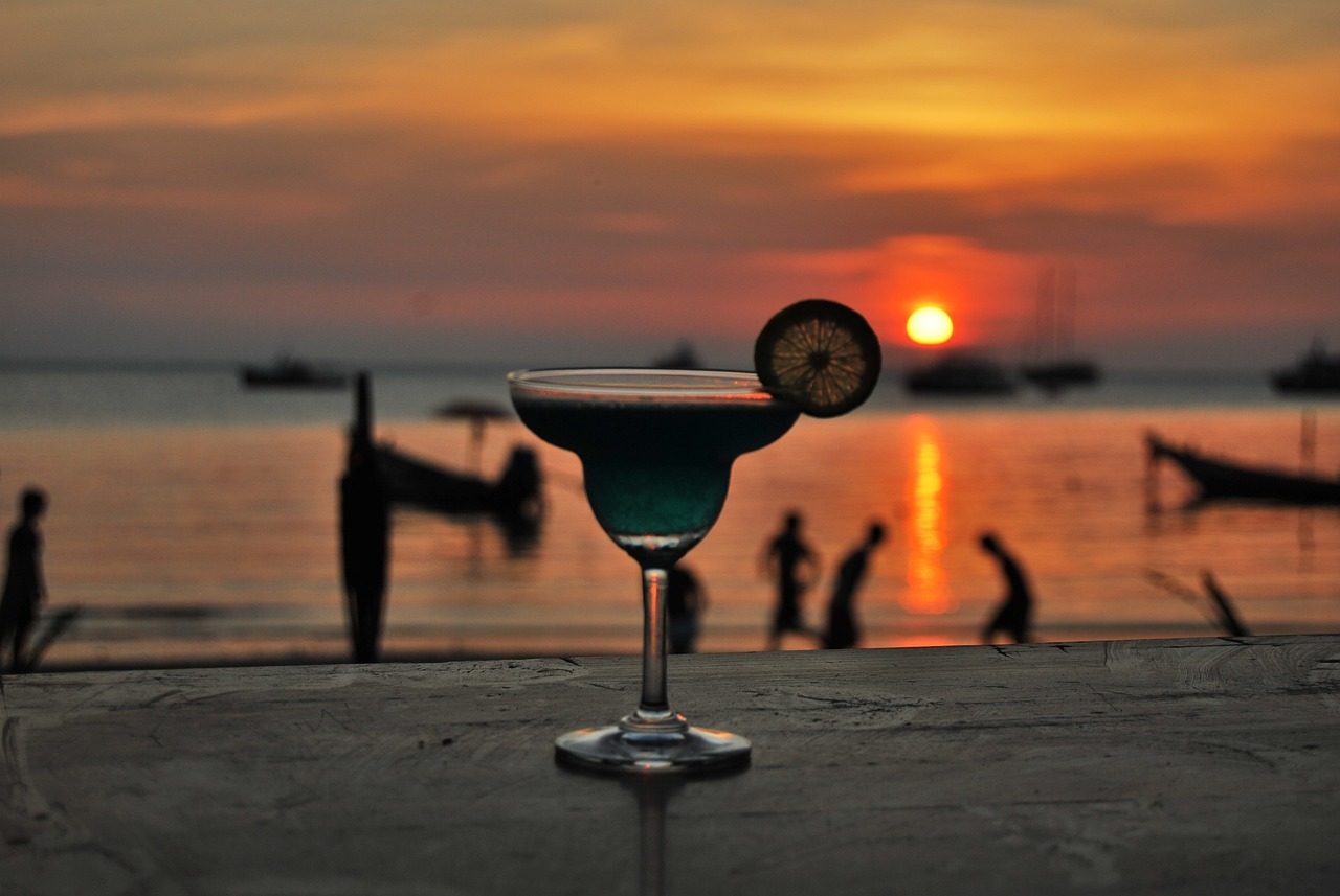 a margarita placed on a deck with a thinly sliced lime overlooks the beach at sunset, as surfers and small fishing boats gather near the shore