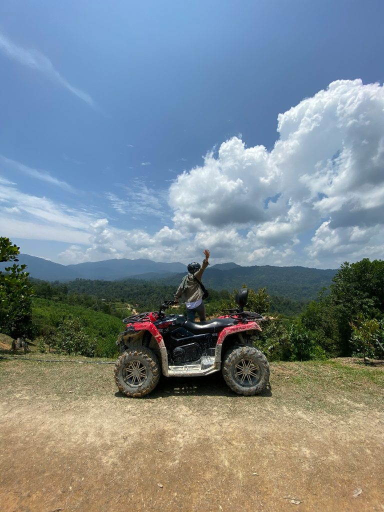A person is standing on an ATV with a hand to the sky, as soft white clouds roll over the hill of rainforests