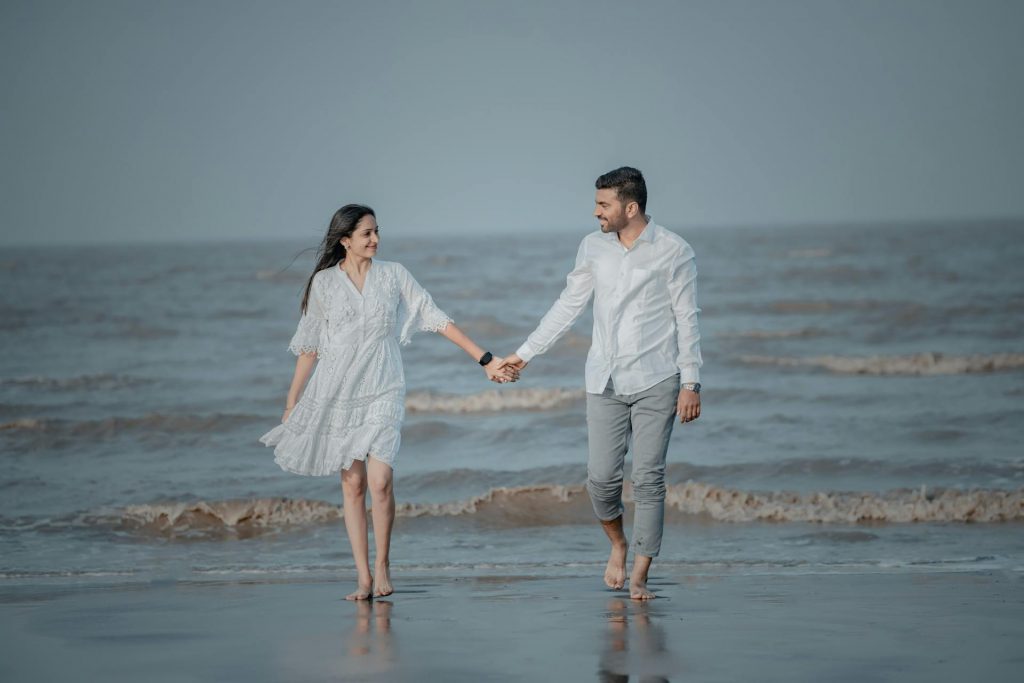 Man and woman holding hands on a beach; woman is wearing a loose white dress while man is wearing a white button-down and gray pants