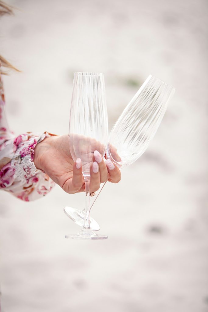 An engaged woman’s manicured hand holding two wine glasses at the beach
